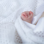 A baby's hand nestled against the soft satin trim of a white brushed cotton cellular blanket